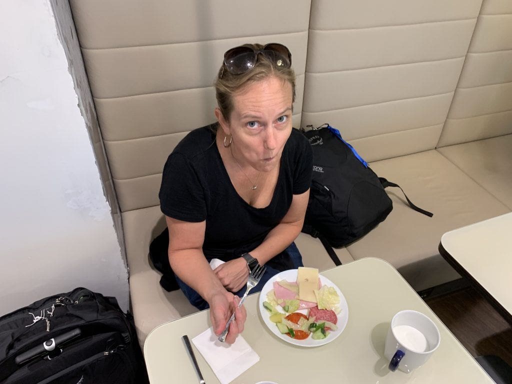 Lisa eating a plate of cold cuts, cheese and vegetables at an airport lounge table, with her carry-on bags beside her