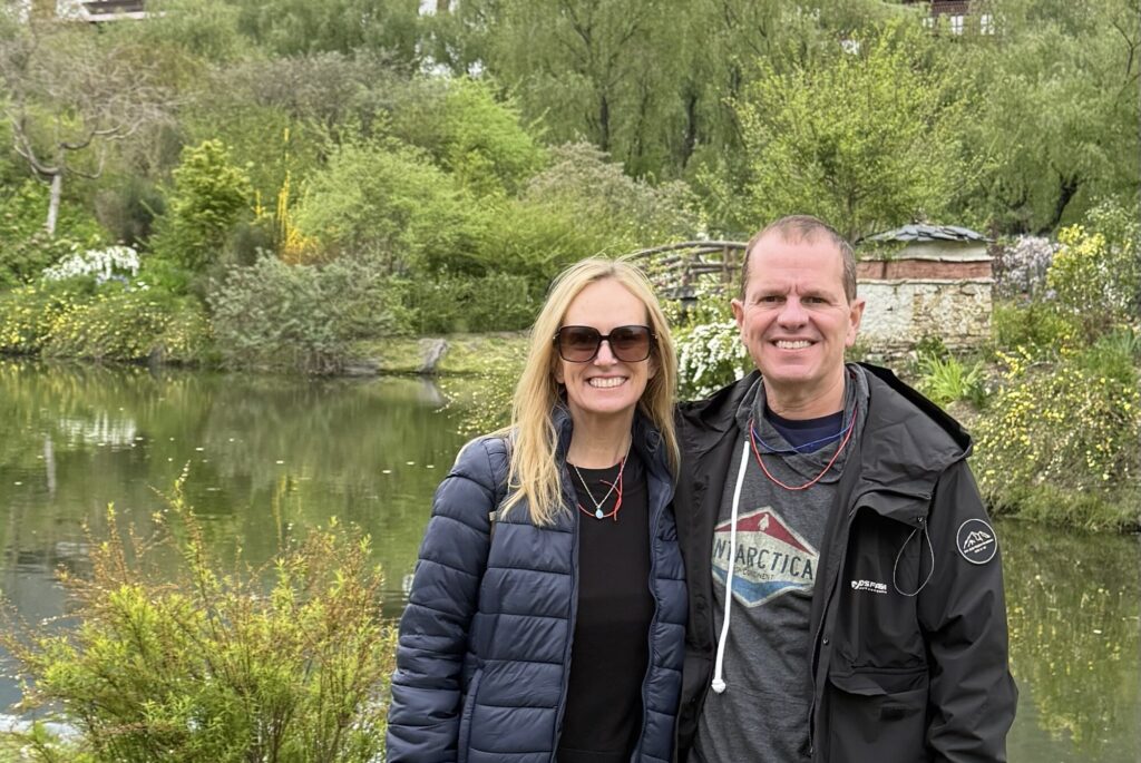 Lisa and Matt standing together beside a pond in Bhutan, with wooded hills behind them