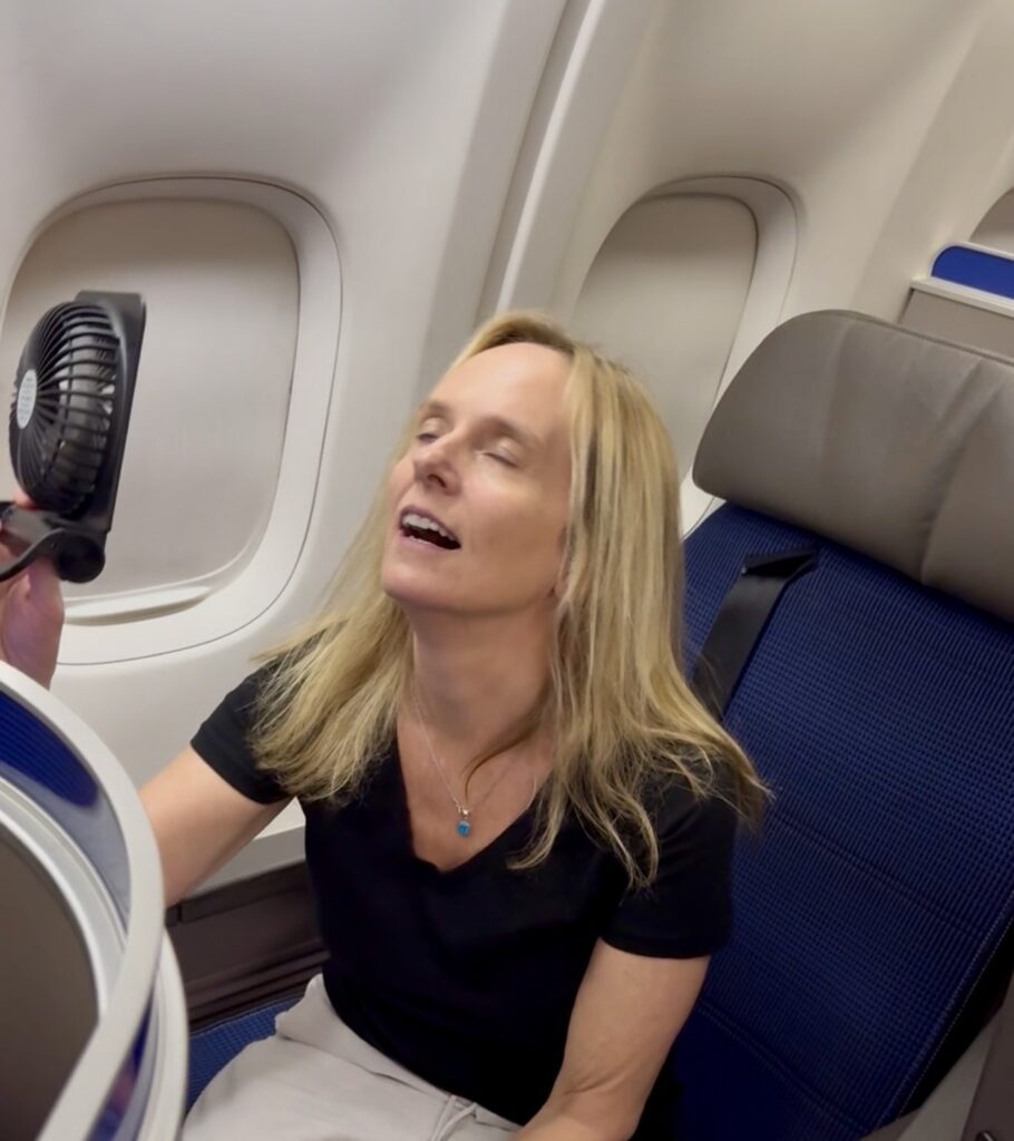 Lisa in an airplane window seat with her eyes closed, cooling off in front of a small handheld fan by the cabin window.
